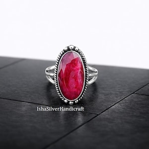 May include: A sterling silver ring with an oval, faceted, ruby-red gemstone. The ring features a detailed rope-like border around the stone and a split shank design. The ring is displayed on a black surface. The text "IshaSilverHandicraft" is visible.