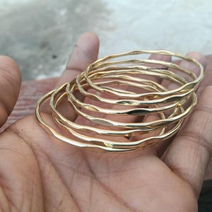 May include: A set of seven gold-colored bangles with a textured, wavy design. The bracelets are stacked on top of each other, creating a layered effect. The jewelry is displayed on a hand.