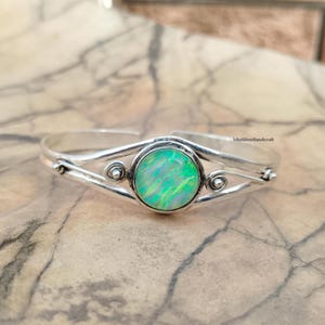 Può includere: Un bracciale a polsino in argento con una grande pietra di opale verde iridescente al centro. Il bracciale ha un design semplice ed elegante con accenti decorativi su entrambi i lati dell'opale. L'opale presenta sfumature di verde e bianco.