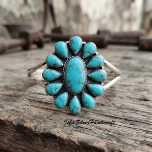 Könnte beinhalten: Ein Silberarmreif mit einem großen, zentralen, ovalen Türkisstein, der von zwölf kleineren, birnenförmigen Türkissteinen in einer blumenartigen Anordnung umgeben ist. Das Metall hat ein oxidiertes Finish und der Text Isha Silver Handicraft ist sichtbar.