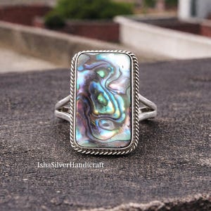 Peut inclure: Bague rectangulaire en argent avec une incrustation de coquillage d'ormeau irisé. La coquille présente un mélange de bleus, de verts et de violets. La bague a une bordure en forme de corde et un design à tige fendue. Le texte "IshaSilverHandicraft" est visible.