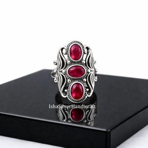 May include: A silver ring with three oval, deep red gemstones in an elaborate, filigree design. The ring has a vintage style, with a polished finish and a detailed setting. The ring is displayed on a black surface.