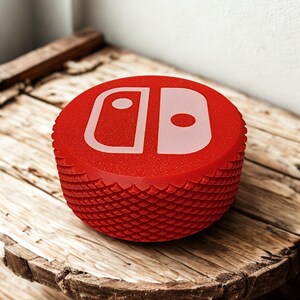 May include: A red, textured controller button with a white graphic of a game console logo. The button is round with a ridged, scale-like pattern around the sides. The button is sitting on a weathered wooden surface.