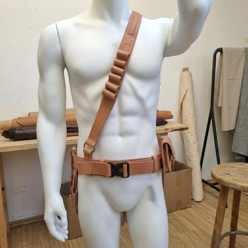 May include: A tan leather bandolier and utility belt set worn on a mannequin. The bandolier features multiple loops for storage, while the belt includes a black plastic side-release buckle and a side pouch for additional gear storage.