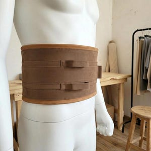 May include: A wide brown suede corset belt featuring two horizontal straps across the front, each secured with a small loop. The belt has a smooth, matte texture and is worn around the waist of a white mannequin, showcasing a structured design.