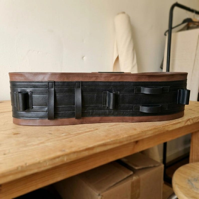 May include: A wide belt featuring a brown leather top and bottom border with a central black leather section. The black section includes multiple vertical leather loops and horizontal stitched detailing across the length of the belt.