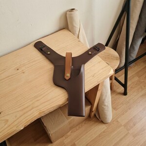 May include: A dark brown leather shoulder holster featuring a tan leather strap with a brass-colored snap button closure. The holster has two adjustable side straps with black metal buckles and a structured, vertical sleeve design for holding an object.