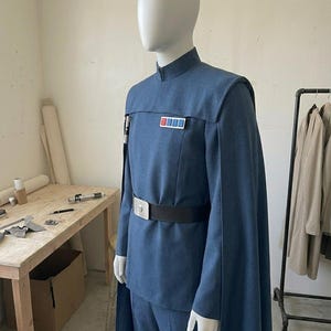 K&ouml;nnte beinhalten: Eine blaue Uniform im Milit&auml;rstil an einer Schaufensterpuppe, bestehend aus einer Tunika mit Stehkragen, passender Hose und einem langen Umhang. Ein schwarzer G&uuml;rtel mit silberner Schnalle wird in der Taille getragen, dazu ein Rangabzeichen.