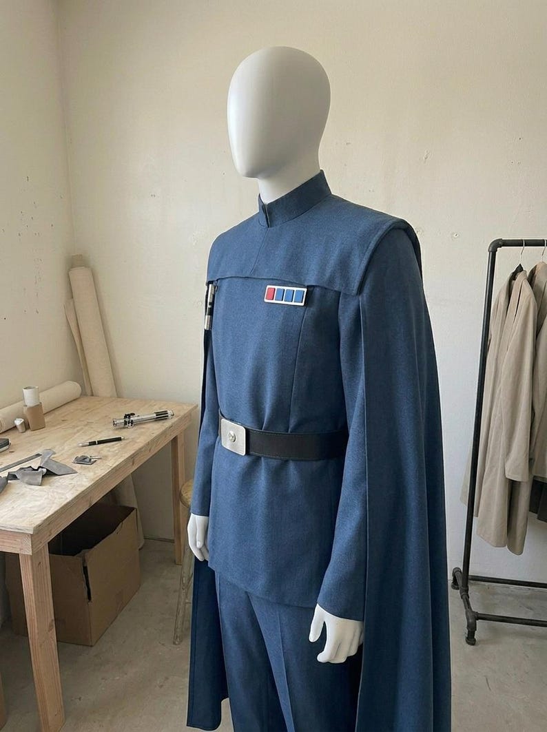 K&ouml;nnte beinhalten: Eine blaue Uniform im Milit&auml;rstil an einer Schaufensterpuppe, bestehend aus einer Tunika mit Stehkragen, passender Hose und einem langen Umhang. Ein schwarzer G&uuml;rtel mit silberner Schnalle wird in der Taille getragen, dazu ein Rangabzeichen.