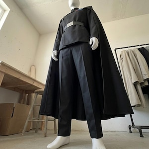 K&ouml;nnte beinhalten: Ein schwarzes Uniformset bestehend aus einer lang&auml;rmeligen Tunika mit Stehkragen, einer passenden Hose und einem bodenlangen Umhang. Die Tunika verf&uuml;gt &uuml;ber eine silberfarbene G&uuml;rtelschnalle und einen kleinen rechteckigen Rangabzeichenbalken.