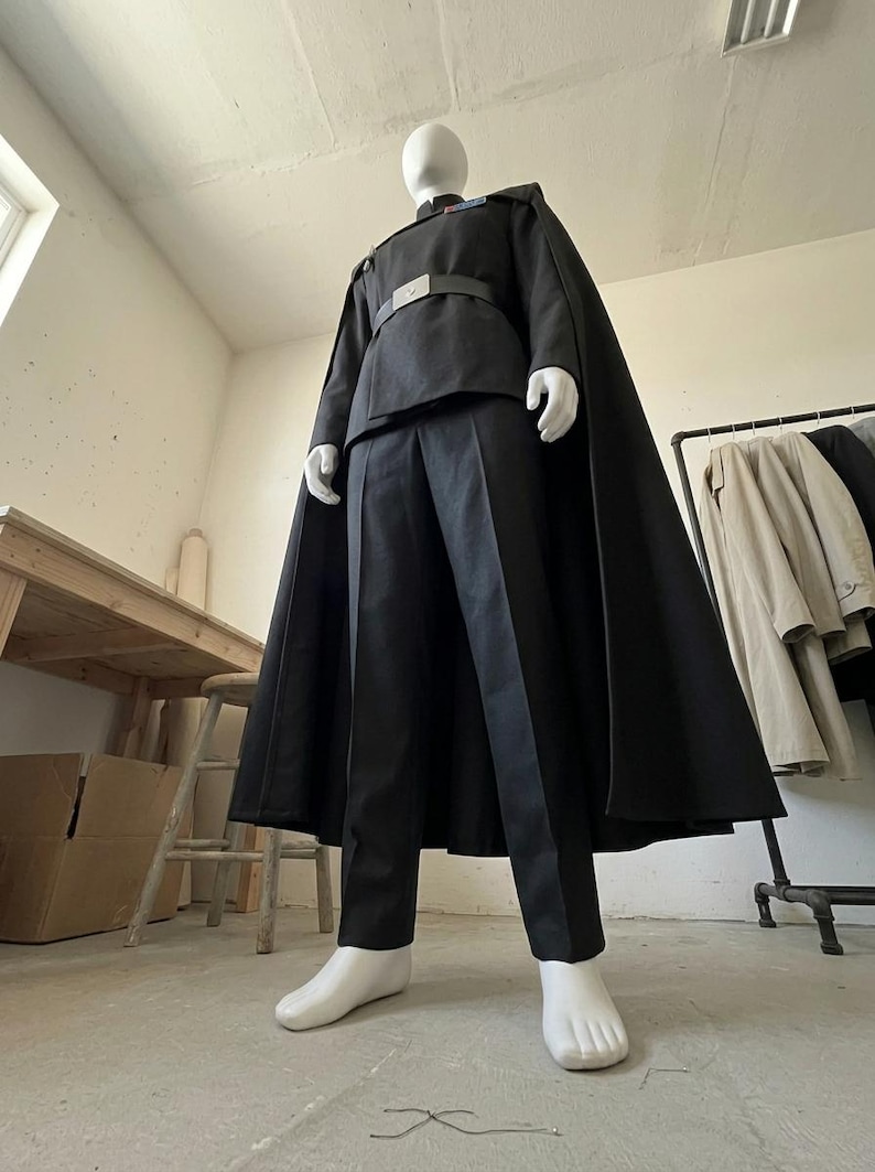 K&ouml;nnte beinhalten: Ein schwarzes Uniformset bestehend aus einer lang&auml;rmeligen Tunika mit Stehkragen, einer passenden Hose und einem bodenlangen Umhang. Die Tunika verf&uuml;gt &uuml;ber eine silberfarbene G&uuml;rtelschnalle und einen kleinen rechteckigen Rangabzeichenbalken.