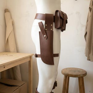 May include: A brown leather utility belt system featuring a waist belt, a large rectangular pouch with a flap closure, a long vertical holster with a silver snap button, and a matching leg strap with a buckle, worn on a white mannequin.