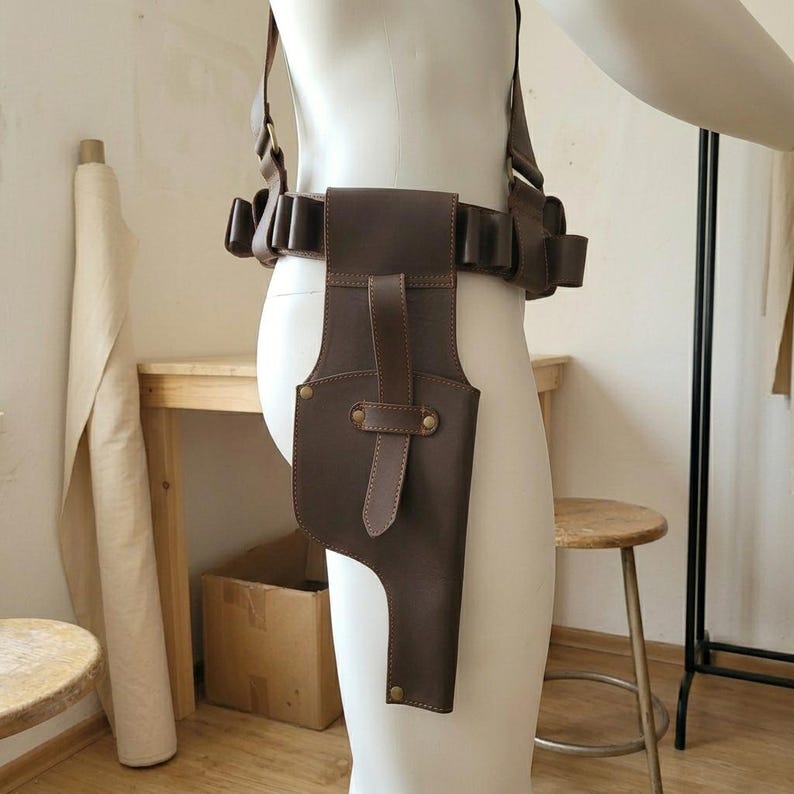 K&ouml;nnte beinhalten: Ein dunkelbraunes Lederholster- und G&uuml;rtelsystem mit einer vertikalen Tasche mit Riemenverschluss, mehreren seitlichen Schlaufen zur Aufbewahrung und verstellbaren Schultergurten, getragen an einer Schaufensterpuppe zur Darstellung.
