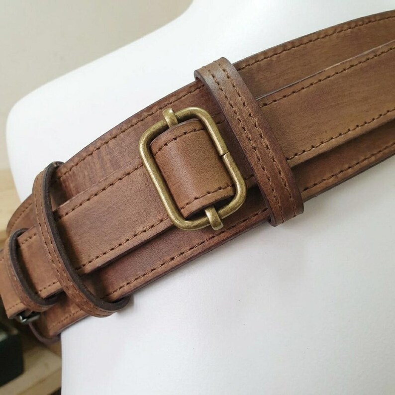 May include: A wide brown leather belt featuring a rectangular antique brass buckle and a matching leather keeper loop. The belt has visible stitched edges and a smooth, matte finish, showcasing a sturdy construction with a classic metal hardware design.