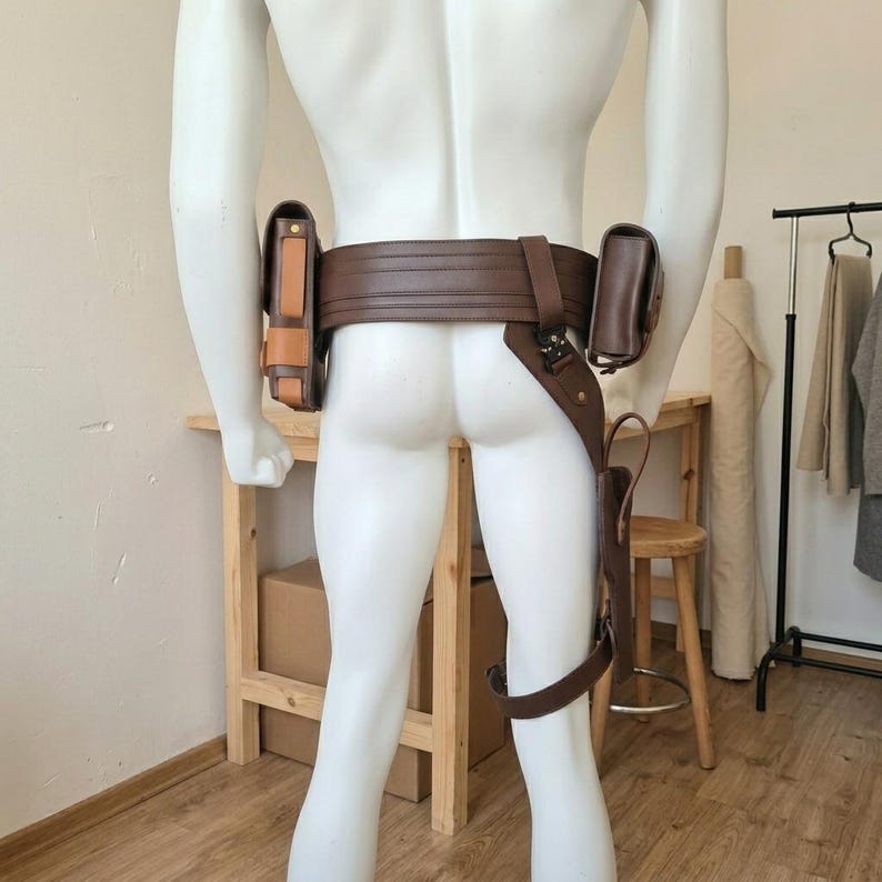 May include: A brown leather utility belt worn on a white mannequin, featuring a wide waistband, two rectangular pouches attached to the sides, and a drop-leg holster component secured with a strap around the thigh area for equipment storage.