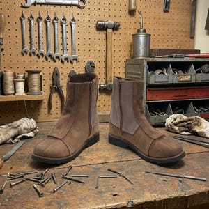 May include: A pair of brown leather boots with a side zipper and a light brown panel. The boots are on a wooden workbench with tools in the background. The boots have a dark brown sole.