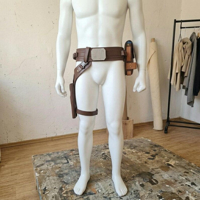 May include: A brown utility belt worn on a white mannequin, featuring a rectangular silver buckle, a drop-leg holster attachment on the right side, and a small rectangular pouch secured to the left side of the waist with metallic hardware.