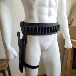 May include: A black leather gun belt featuring a series of integrated cartridge loops along the waist and a matching holster attached to a leg strap. The holster is positioned on the right side, secured with a vertical strap and a horizontal thigh band.