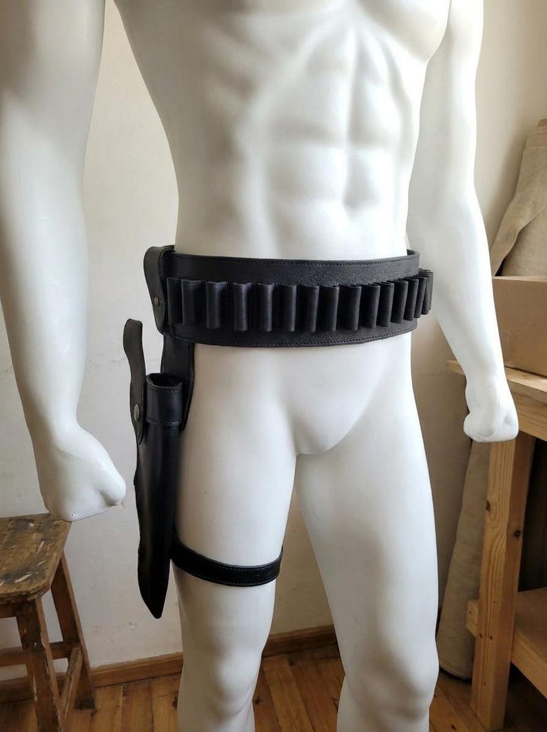 May include: A black leather gun belt featuring a series of integrated cartridge loops along the waist and a matching holster attached to a leg strap. The holster is positioned on the right side, secured with a vertical strap and a horizontal thigh band.