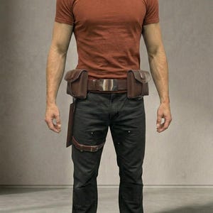 Op de afbeelding: Een roestkleurig T-shirt met korte mouwen gecombineerd met een zwarte broek en een bruine leren gereedschapsriem. De riem is voorzien van twee zijtassen, een centrale metalen gesp en een bruine leren beenband aan een holster.