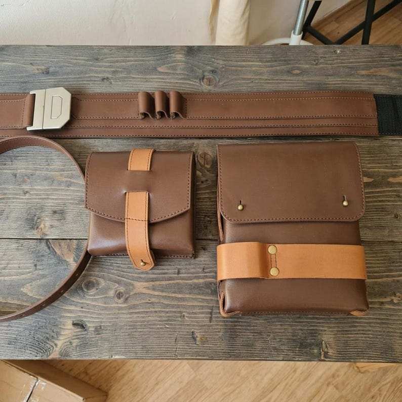 May include: A brown leather utility belt featuring a silver-toned buckle and three small loops, accompanied by two separate brown leather pouches. One pouch is square with a tan leather strap, and the other is rectangular with a tan leather band.