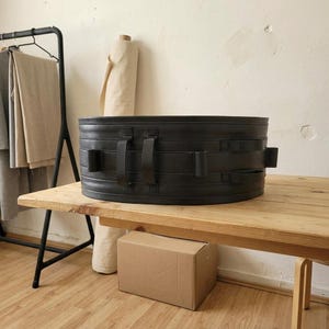 May include: A wide, cylindrical black storage container featuring horizontal stitched detailing and multiple vertical fabric handles attached to the exterior. The container has a structured, solid form with a smooth finish and is positioned on a wooden surface.