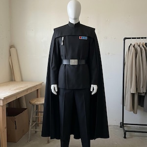 K&ouml;nnte beinhalten: Eine schwarze Uniform im Milit&auml;rstil mit einer Tunika mit hohem Kragen, passender Hose und einem langen Umhang. Die Tunika verf&uuml;gt &uuml;ber ein rechteckiges Rangabzeichen mit roten und blauen Quadraten auf der Brust, ein silbernes zylindrisches Detail und einen schwarzen G&uuml;rtel.
