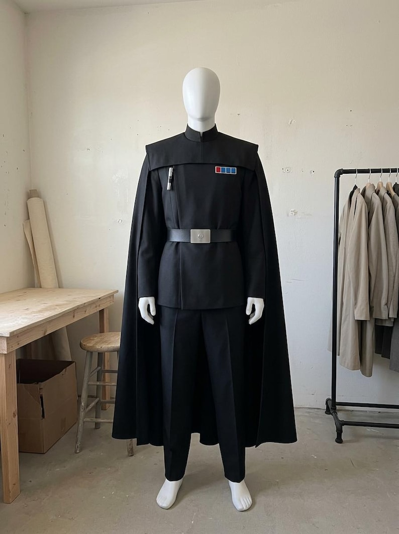 K&ouml;nnte beinhalten: Eine schwarze Uniform im Milit&auml;rstil mit einer Tunika mit hohem Kragen, passender Hose und einem langen Umhang. Die Tunika verf&uuml;gt &uuml;ber ein rechteckiges Rangabzeichen mit roten und blauen Quadraten auf der Brust, ein silbernes zylindrisches Detail und einen schwarzen G&uuml;rtel.