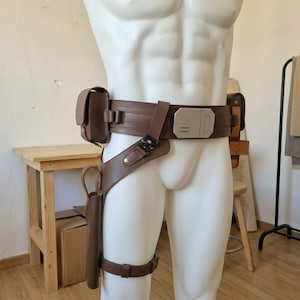 May include: A brown utility belt featuring a rectangular metallic buckle with engraved lines, a side pouch, and a drop-leg holster with a thigh strap. The belt is constructed from leather-like material with metallic hardware and a black tactical buckle.