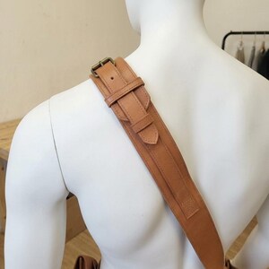 May include: A tan leather shoulder strap featuring an adjustable buckle mechanism with a metal pin and a sliding keeper loop. The strap is wide and flat, constructed with visible stitching along the edges, and rests across the shoulder.