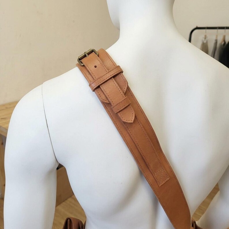 May include: A tan leather shoulder strap featuring an adjustable buckle mechanism with a metal pin and a sliding keeper loop. The strap is wide and flat, constructed with visible stitching along the edges, and rests across the shoulder.