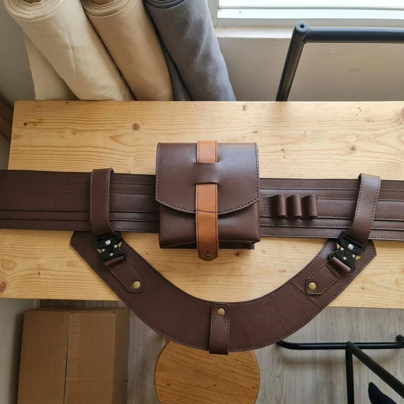 May include: A dark brown leather utility belt featuring a central rectangular pouch with a tan leather strap closure. The belt includes a curved lower leather section attached with black metal buckles and brass-colored rivets, plus three small loops.