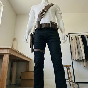Pode incluir: Uma t-shirt henley branca de manga curta e cal&ccedil;as de ganga azul-escuras usadas com um cinto utilit&aacute;rio e uma al&ccedil;a de ombro em pele castanha. O cinto apresenta v&aacute;rias bolsas, um coldre e uma al&ccedil;a com detalhes cil&iacute;ndricos met&aacute;licos.