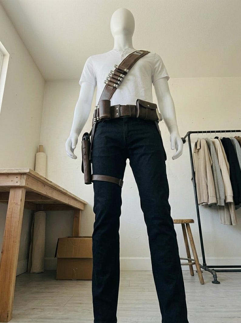 Pode incluir: Uma t-shirt henley branca de manga curta e cal&ccedil;as de ganga azul-escuras usadas com um cinto utilit&aacute;rio e uma al&ccedil;a de ombro em pele castanha. O cinto apresenta v&aacute;rias bolsas, um coldre e uma al&ccedil;a com detalhes cil&iacute;ndricos met&aacute;licos.