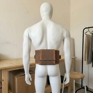 May include: A brown canvas waist bag with a rectangular flap closure and side attachment loops. The bag is secured around the waist of a white mannequin, featuring a structured design with visible stitching and a flat, horizontal orientation.