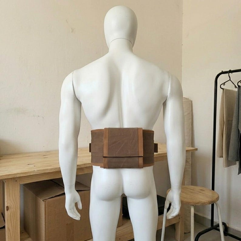 May include: A brown canvas waist bag with a rectangular flap closure and side attachment loops. The bag is secured around the waist of a white mannequin, featuring a structured design with visible stitching and a flat, horizontal orientation.