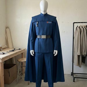 K&ouml;nnte beinhalten: Eine blaue Uniform im Milit&auml;rstil, bestehend aus einer Tunika mit Stehkragen, einem passenden Umhang, einer geraden Hose und einem schwarzen G&uuml;rtel mit silberner Schnalle. Die Tunika hat ein Rangabzeichen mit roten und blauen Quadraten.