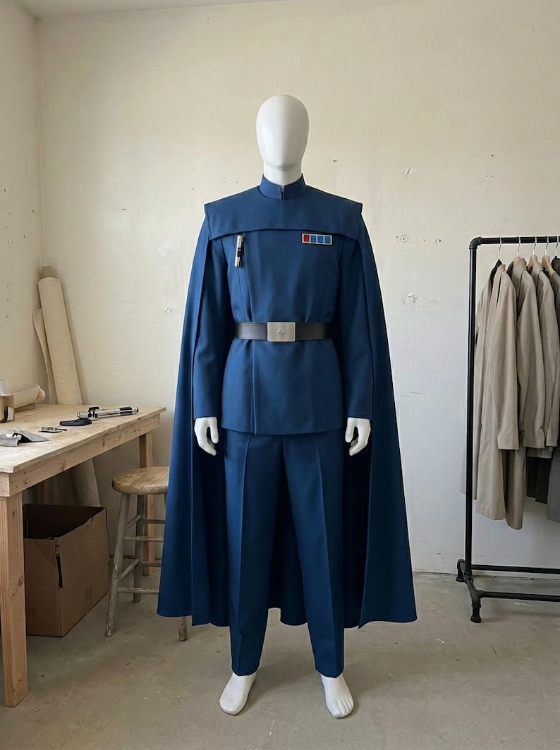 K&ouml;nnte beinhalten: Eine blaue Uniform im Milit&auml;rstil, bestehend aus einer Tunika mit Stehkragen, einem passenden Umhang, einer geraden Hose und einem schwarzen G&uuml;rtel mit silberner Schnalle. Die Tunika hat ein Rangabzeichen mit roten und blauen Quadraten.