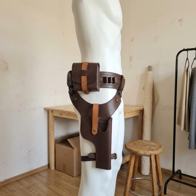 May include: A dark brown leather utility belt featuring a matching holster and a rectangular pouch. The belt includes a lighter tan leather strap detail on the pouch and holster, secured with metal snaps, and three small loops on the side.