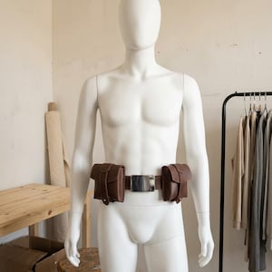 May include: A brown leather utility belt featuring two matching pouches on either side and a large, rectangular, polished silver-tone metal buckle at the centre. The belt is worn around the waist of a white, featureless, abstract mannequin torso.
