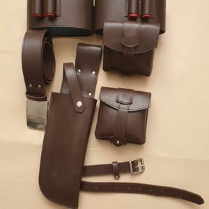 Op de afbeelding: Een set donkerbruine lederen uitrusting bestaande uit twee brede riemen met rode patroonhouders, twee vierkante tasjes met klepsluiting, een lange holster met een zilveren drukknoop en een dunne verstelbare riem met een zilveren gesp.