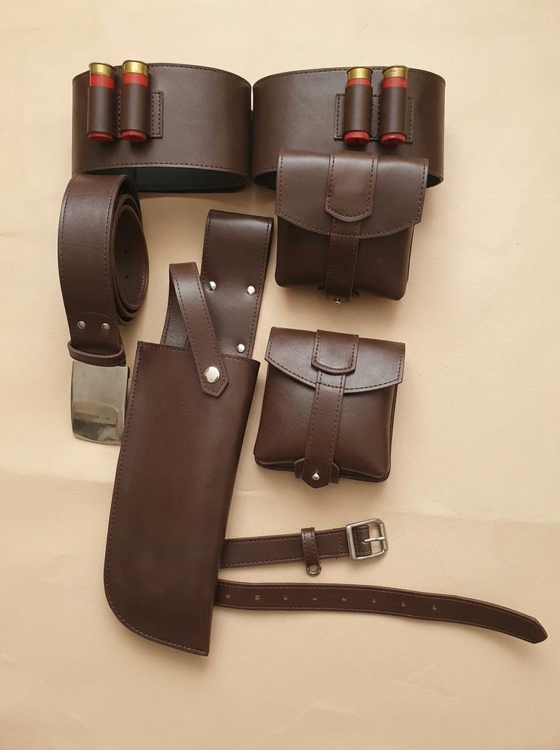 Op de afbeelding: Een set donkerbruine lederen uitrusting bestaande uit twee brede riemen met rode patroonhouders, twee vierkante tasjes met klepsluiting, een lange holster met een zilveren drukknoop en een dunne verstelbare riem met een zilveren gesp.