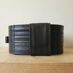 May include: A wide black leather belt featuring horizontal stitched panels and a smooth, vertical central overlay piece. The belt has a structured, multi-layered appearance with clean, straight seams running across the length of the dark material.