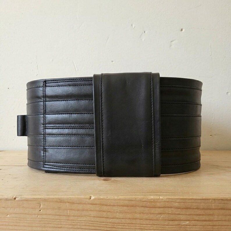 May include: A wide black leather belt featuring horizontal stitched panels and a smooth, vertical central overlay piece. The belt has a structured, multi-layered appearance with clean, straight seams running across the length of the dark material.
