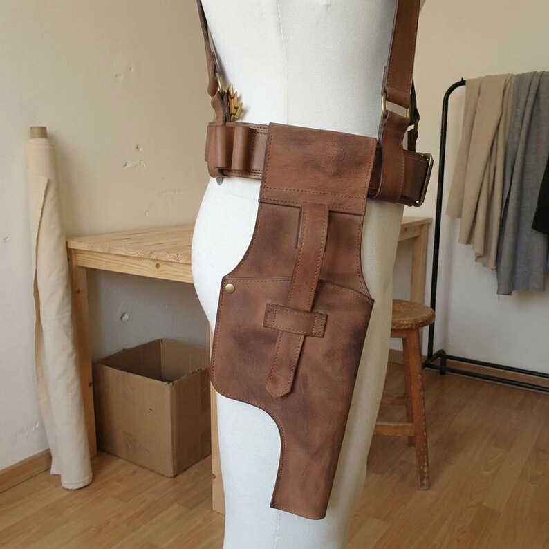 May include: A brown leather holster attached to a wide belt with brass hardware. The holster features a vertical strap closure and a contoured shape, worn around the waist and supported by shoulder straps on a white mannequin torso.