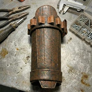 May include: A cylindrical, brown leather tool holder with multiple small pockets around the top. The holder has a stitched design and a strap. Tools, including pliers and a screwdriver, are visible in the background.
