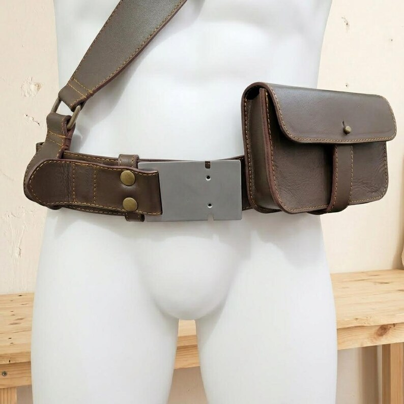 K&ouml;nnte beinhalten: Ein dunkelbrauner Leder-G&uuml;rtel mit einer rechteckigen, silberfarbenen Metallschnalle, einer passenden Ledertasche mit Klappverschluss und einem breiten Schulterriemen, der mit bronzefarbenen Metallbeschl&auml;gen und Nieten befestigt ist.
