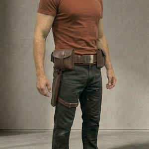 Op de afbeelding: Een roestkleurig T-shirt met korte mouwen gecombineerd met een donkergrijze broek. Een bruine leren utility-riem heeft een grote zilveren gesp, twee bevestigde tasjes en een holster voor aan het been met een riem om de dij.