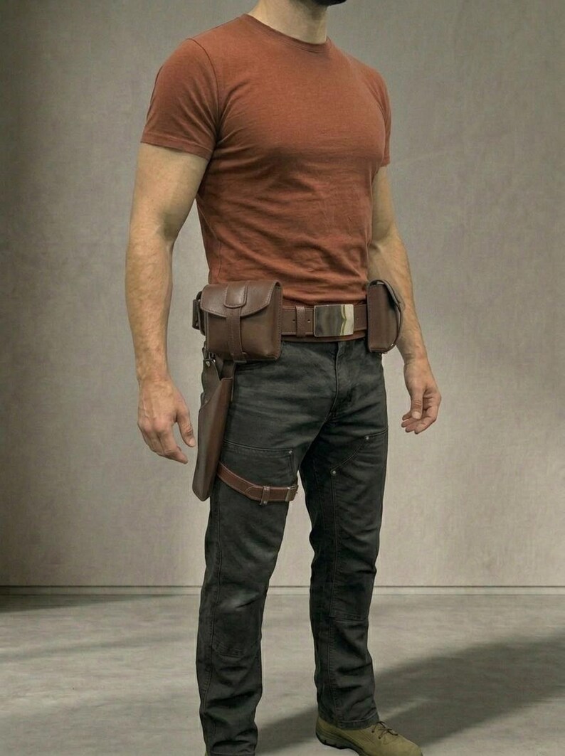 Op de afbeelding: Een roestkleurig T-shirt met korte mouwen gecombineerd met een donkergrijze broek. Een bruine leren utility-riem heeft een grote zilveren gesp, twee bevestigde tasjes en een holster voor aan het been met een riem om de dij.