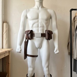 May include: A brown leather utility belt set featuring two side pouches, a central rectangular buckle, a long holster attachment on the right side with a thigh strap, and two matching brown leather leg guards worn on the lower thighs.